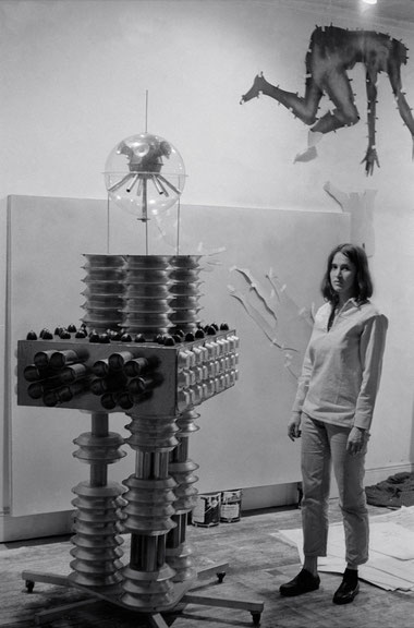 Kiki Kogelnik und ihre Skulptur <i>Lover Boy</i> (1963, Mischtechnik mit Kuchen­ und Muffinformen sowie Zigarrenhülsen, 213 × 70 × 120 cm) in ihrem Atelier in New York, 1965, Fotografin unbekannt